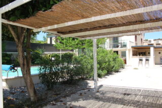 Terrasse ombragée sous pergola en canisse avec vue sur le jardin, la piscine turquoise et la maison du Mas du Camisard à Ribaute-les-Tavernes (Gard)