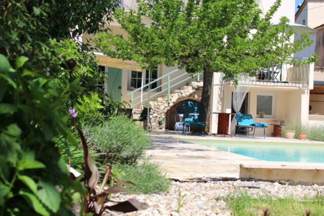 Vue du Mas du Camisard depuis le jardin, avec l’allée en graviers, la piscine, la façade principale et les arbres méditerranéens